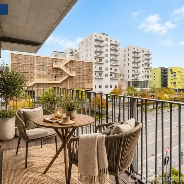En balkon med et bord og to stole indeholder tændte fyrfadslys, krus og planter. Moderne bygninger og træer ses i baggrunden. Tekst: "Visualisering".