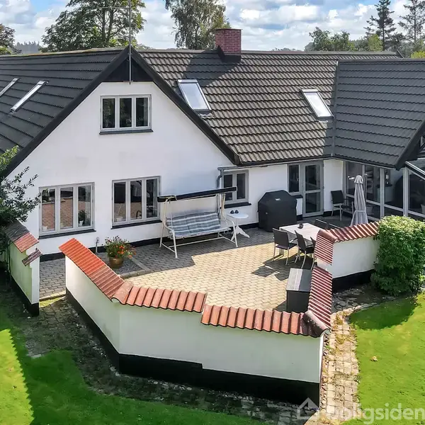 Hus med sort tag og hvide vægge; har en terrasse med møbler og grill; omgivet af en velplejet have med træer og buske.