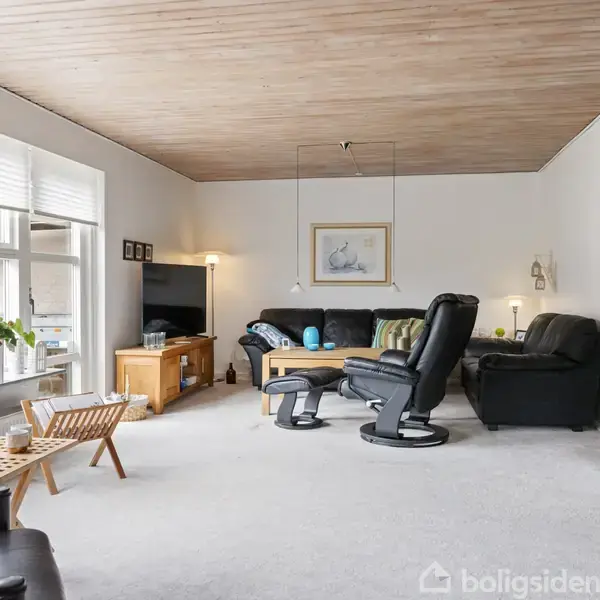 Stueindretning med sorte lænestole og sofa, placeret omkring et sofabord. TV i hjørnet, vindue til venstre med planter. Lys trængende ind. Buet døråbning i baggrunden.