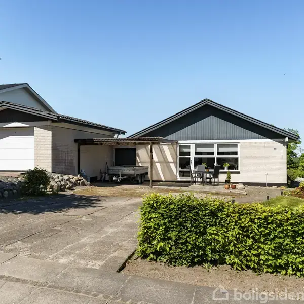 Et enkelt enfamiliehus med garage og fladt tag står centralt. En gårdsplads med flisebelægning foran, omgivet af grønne buske og træer i solrigt vejr.