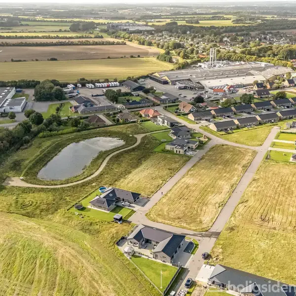 Landlige huse ligger spredt omkring en central dam, omgivet af åbne marker og et industriområde i baggrunden. Små veje forbinder boligerne med det omkringliggende landskab.