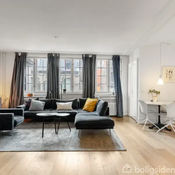 Stue med mørkegrå sofa og sofabord, placeret på et lyst trægulv. Store vinduer med lange gardiner i baggrunden. Rundt spisebord med to hvide stole og hængende lampe i højre side.