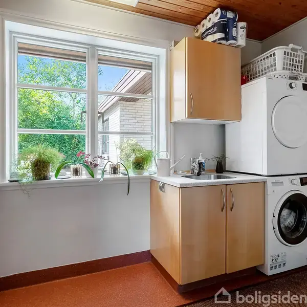 Vaskemaskine og tørretumbler stablet i et bryggers, omgivet af trækabejskabe og vask under et vindue med grøn udsigt. Planter pryder vindueskarmen, mens kurve står øverst.