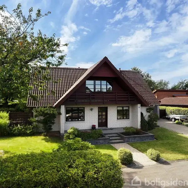 Et to-etagers hus med rødt tegltag og trædetaljer står midt i en velplejet have. En flisebelagt sti fører til hoveddøren. Himlen er blå med lette skyer.