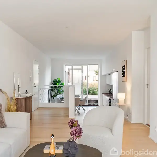 En lys stue med hvide vægge og en hvid sofa. Et rundt sofabord med blomster står i midten. Store vinduer i baggrunden giver naturligt lys.