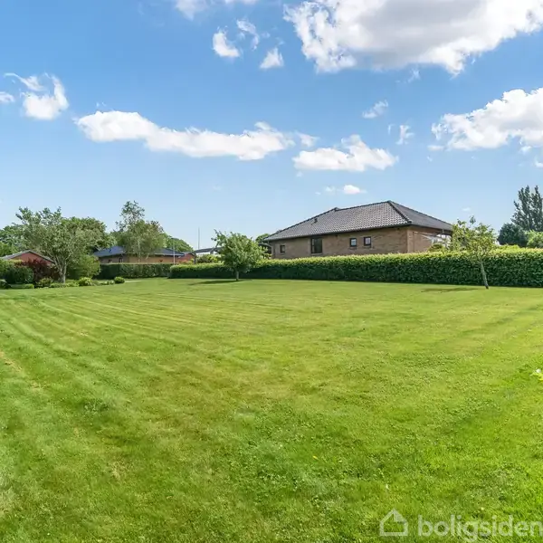 En velplejet græsplæne strækker sig foran et moderne hus, omgivet af grønne buske under en blå himmel med spredte skyer.