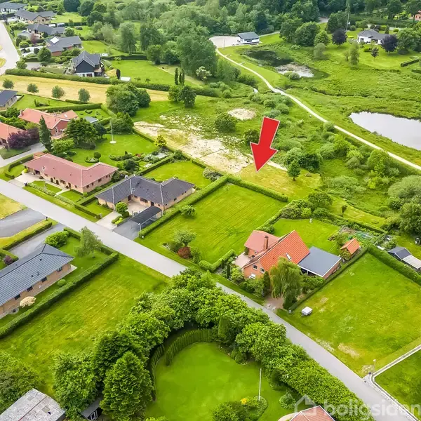 Et parcelhus med rødt tag ses fra luften. Det er omgivet af en grøn have og markeret med en rød pil. Omgivelserne inkluderer andre huse og et grønt område med en sø.