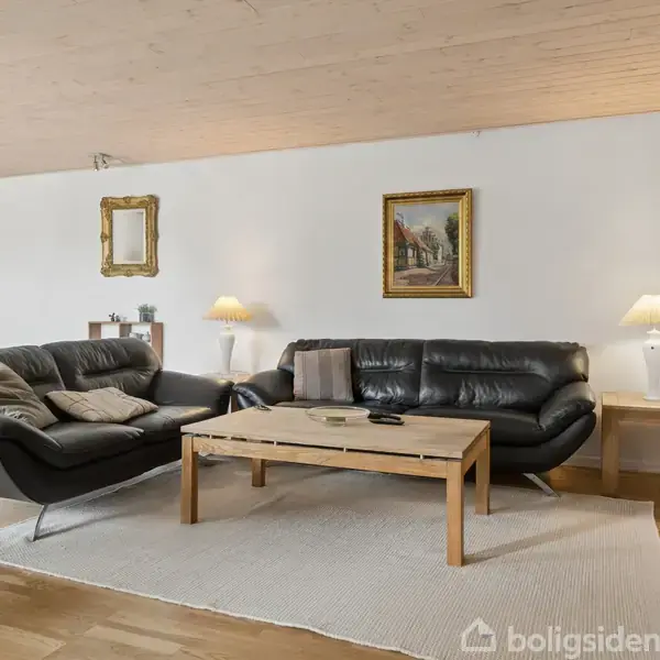 To sorte sofaer står på et lyst tæppe omkring et rektangulært træbord. Baggrunden har en hvid væg med indrammet kunst. En lampe lyser rummet.