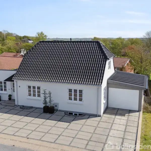 Hvidt hus med sort tag står stille på en stor flisebelagt indkørsel; omgivet af frodig, grøn natur med flere huse i nærheden.