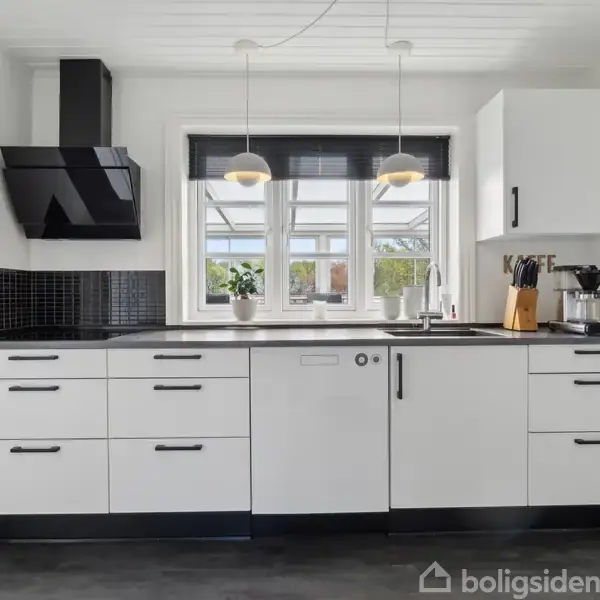Et moderne køkken med hvide skabe og sorte håndtag, en sort emhætte og to hængende lamper. Et vindue med planter ved vasken kigger ud til en have.