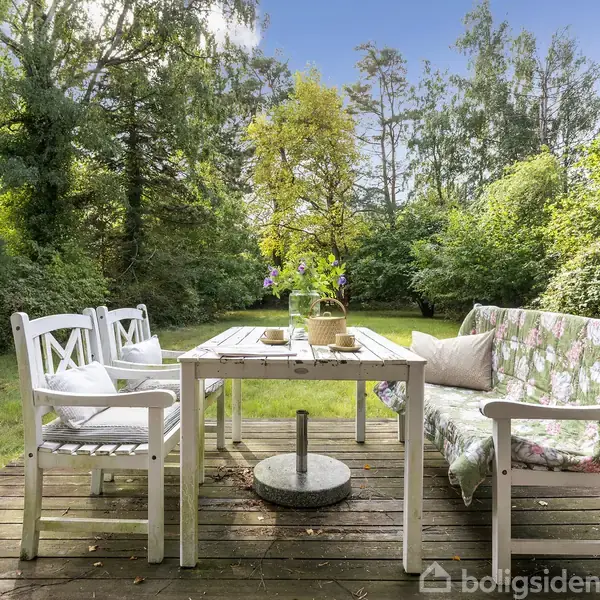 Et træbord med blomster i vase på står på en træterrasse. Omkring bordet er der hvide stole og en sofa med blomstret betræk. Haven er omgivet af frodige træer.