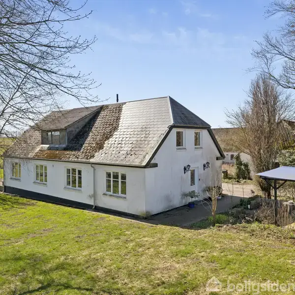 Et hvidt, landligt hus med skråt tag står i en grøn have. Et lyst overdækket siddeområde ses bagved. Træer omkranser ejendommen på en solrig dag.