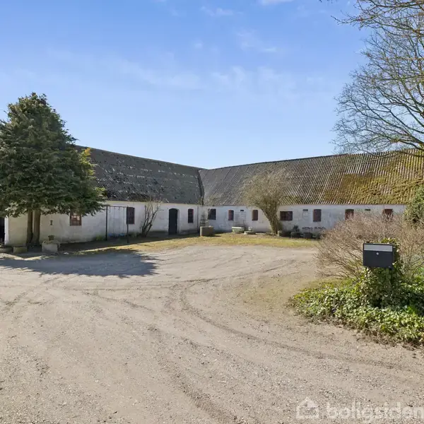 En firlænget gård med hvidkalkede mure står stille under en blå himmel. Træer skaber skygge og grusvejen løber gennem gårdspladsen. En postkasse står ved siden af buske.
