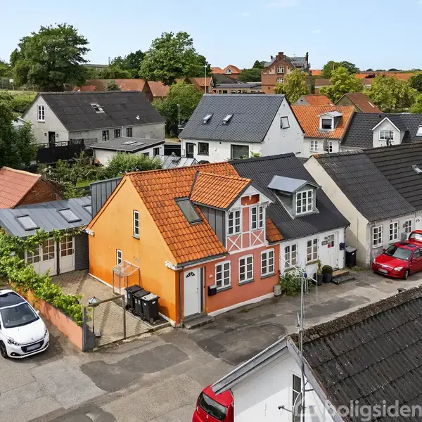 Et orange hus med hvide detaljer står midt i en række af farverige huse. Foran parkerer to biler, og grønne træer omkranser den stille gade i et boligområde.