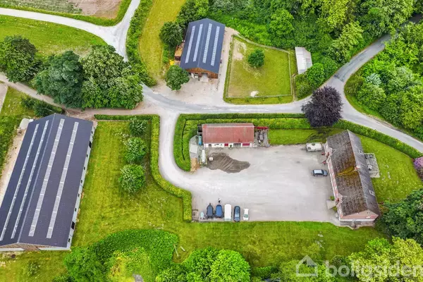 Tre bygninger med sorte tage omgiver en central gårdsplads; en lang bygning til venstre og to mindre i midten og højre. Bygningerne er omgivet af græs og træer.