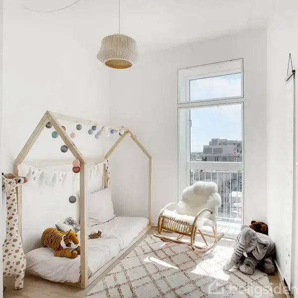 Et minimalistisk børneværelse med en træseng formet som et hus, dekoreret med guirlander. Ved siden af ses en gyngestol i sollys fra et stort vindue med byudsigt.