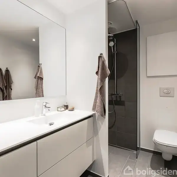 Badeværelse med hvidt håndvaskskab og stor spejl. Til venstre hænger beige håndklæder. I højre hjørne ses en bruseniche og toilet. Miljøet er lyst og moderne.