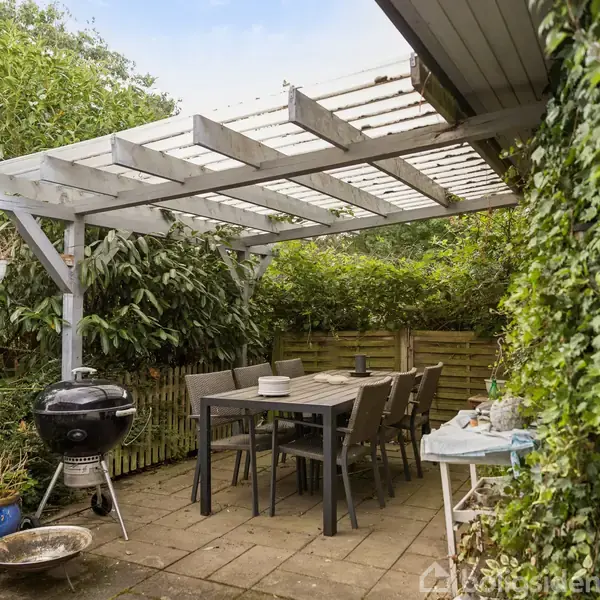 Terrasse med spisebord og seks stole under en pergola. En kuglegrill står ved siden af bordet. Omgivet af frodige grønne planter og et plankeværk.