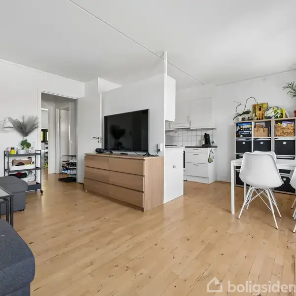 Et lyst stueområde med trælaminatgulv. Fjernsynet står på en skænk, omgivet af sofaer. Til højre er der et køkkenhjørne og to hvide stole ved et lille bord.