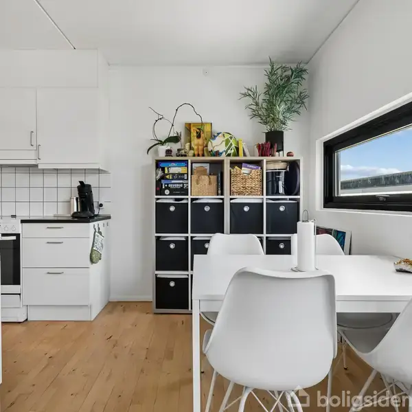 Et moderne køkken med hvide skabe og komfur. Et bord med fire hvide stole står ved vinduet. En reol med kurve og planter står i hjørnet.