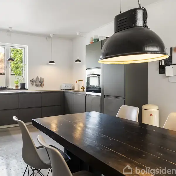 Et moderne køkken med mørke skabe og en central spisebord med hvide stole. En stor sort lampe hænger over bordet. Lys strømmer ind fra et vindue mod venstre.