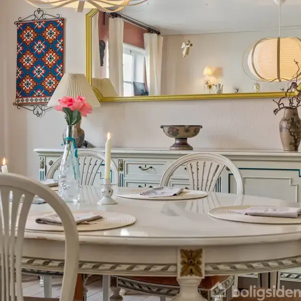 Rundt spisebord med hvide stole står dækket med tallerkener og stearinlys i en hyggelig spisestue. En stor guldspejl hænger over et hvidt skab med dekorationer og en vase med blomster.