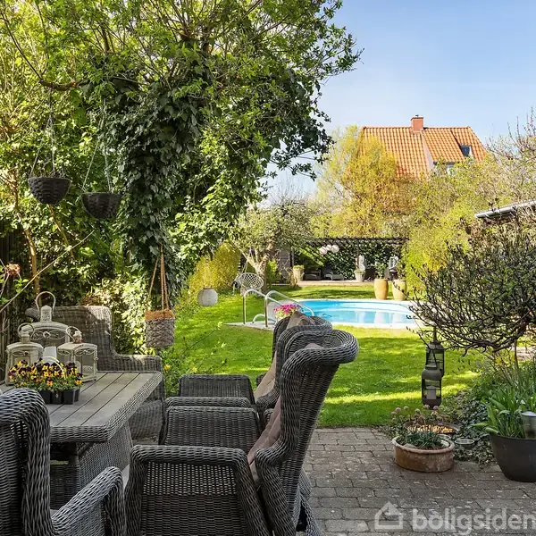 Terrassemøbler står omkring et bord med blomsterdekorationer, omgivet af en grøn have med træer og en swimmingpool i baggrunden. Haven er tæt og frodig under en klar blå himmel.