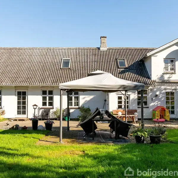 En hvid villa med gråt tag har en solbeskinnet terrasse med et telt, borde og stole. Haven omkring er grøn og velplejet med forskellige planter og en legetøjsrutsjebane.