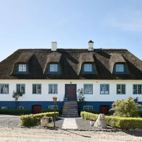 Et stråtækt hus står stille i en have, omgivet af buske og træer. En trappe fører til hoveddøren, mens grus og planter skaber en indbydende forhave.
