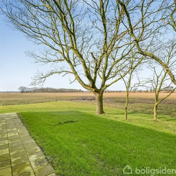 Husfacade med terrasse på venstre side; store træer står i græsplæne; vidtstrakt mark og himmel danner baggrund.