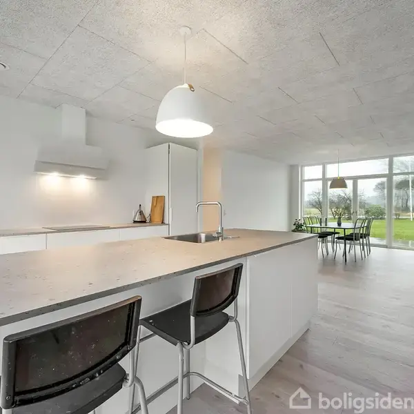 Et moderne køkken med en stor ø midt i rummet og to sorte barstole. Køkkenet har en hvid lampe og tilstødende spiseområde med udsigt til en have udenfor.