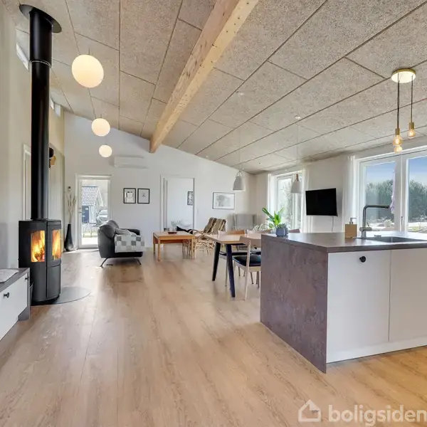 Pejs kører ved siden af moderne køkkenø i et lyst, rummeligt stueområde med spiseplads og sofahjørne. Store vinduer giver udsigt til haven. Text: "boligsiden".