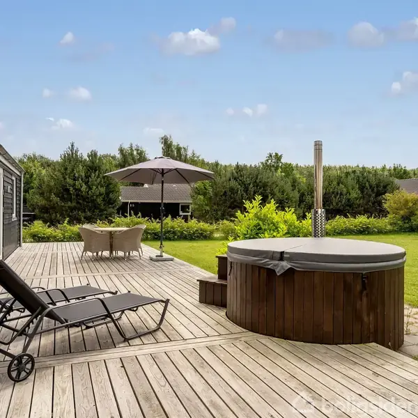 Et trædæk i en have med to liggestole og et rundt vildmarksbad. I baggrunden står et bord med parasol, omgivet af grønne buske og huse.