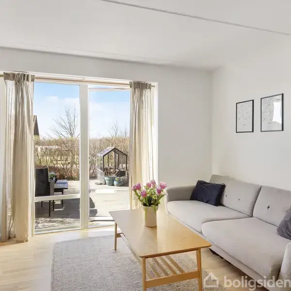 Stue med en beige sofa, sofabord og blomster, vendt mod en glasdør, der fører ud til en terrasse med havemøbler. Lysindfald fra dørens vindue oplyser rummet.