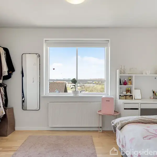 Værelset har en seng med blomsterdyne til højre, en skrivebord ved siden af med hylde, og en stor vindue i midten med udsigt over huse. Garderobe og spejl er til venstre.