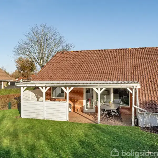 Hus med rødbrun tegltag og en lille overdækket terrasse med bord og stole. Græsplæne og hæk omkranser huset. Klart vejr og flere huse i baggrunden.