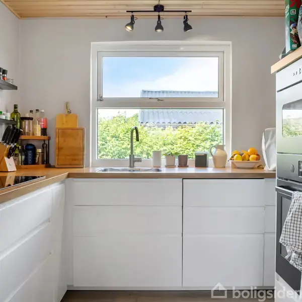 Hvidt køkken med moderne design har hvidlakerede skabe og integrerede apparater. Et vindue over vasken giver udsigt til grøn beplantning udenfor. En skål med appelsiner står på bordpladen.