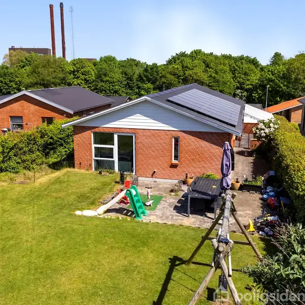 Rødt murstenshus med solpaneler står i en grøn have. Haven har en terrasse med møbler og legetøj. Omgivet af andre huse og fyldige buske under en blå himmel.