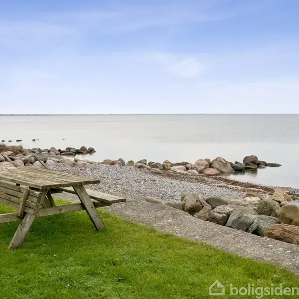 Bordbænkesæt står på en græsplæne tæt på stranden. Kystlinjen har en stensætning, og havet strækker sig ud i horisonten under en klar himmel.