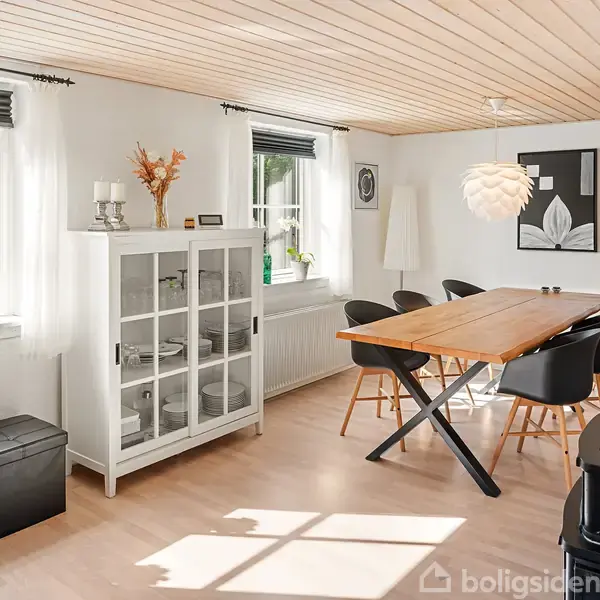 Et træbord med sorte stole står i en lys spisestue. Til venstre er et hvidt vitrineskab med glasdøre, dekoreret med en vase og lysestager.