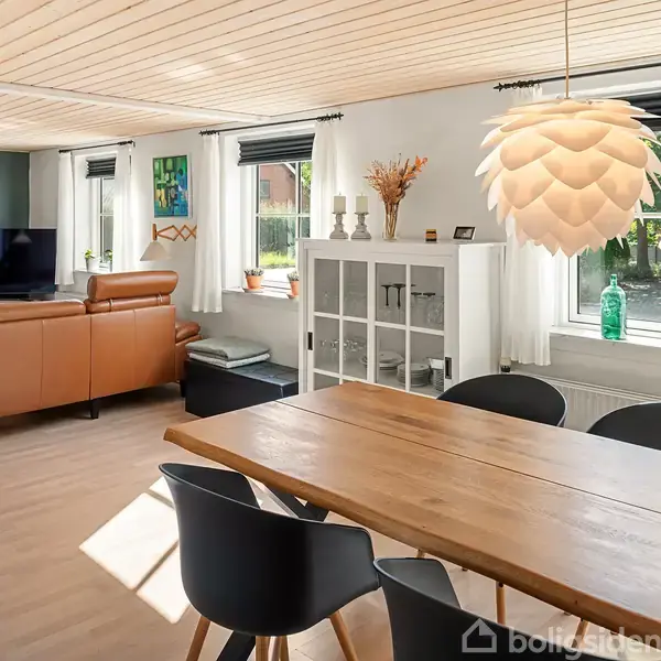 Et lyst og rummeligt stueområde indeholder et spisebord med sorte stole, en stor gulvlampe, og en sofagruppe med fjernsyn. Store vinduer giver udsigt til en grøn have.