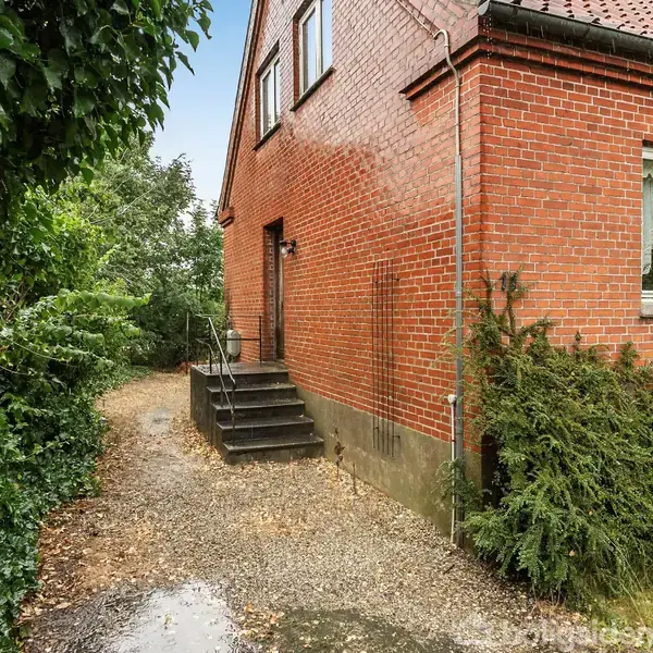 Rødt murstenshus med trappe ved indgangen, omkranset af grønne buske. Indgangen leder til en smal sti. Vinduerne har hvide rammer. Huset har et rødt tegltag.