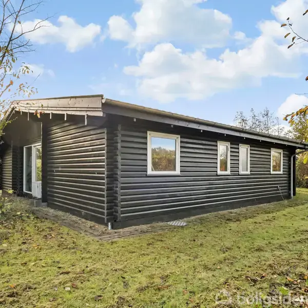 Et sort træhus står stille i en frodig have med græsplæne og træer omkring. Sollys skinner svagt på husets facade.