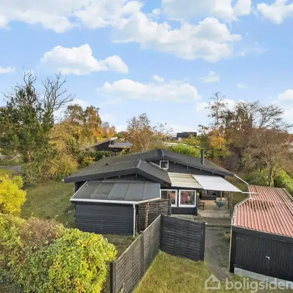 Et sommerhus med sort tag og vægge står i en grøn have omkranset af buske. Baggrunden viser flere huse og blå himmel med skyer.