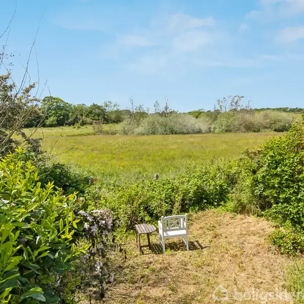 En hvid stol og et lille bord står på kortklippet græs ved siden af buske. Foran strækker sig en stor grøn mark under en blå himmel.