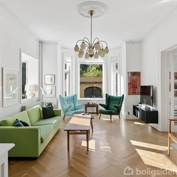 Stue med grøn sofa og to turkise stole omkring et sofabord. Store vinduer lader sollys ind, og en moderne lysekrone hænger fra loftet. Lyse trægulve.