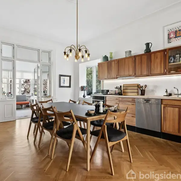 Spisebord med træstole står i et køkken. Køkkenet er udstyret med træoverskabe, opvaskemaskine og væghængte knager. En moderne lysekrone hænger over bordet.