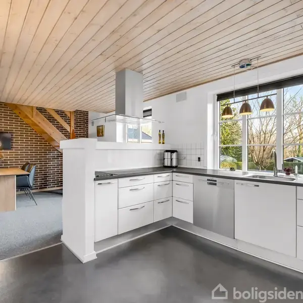 Køkken med hvide skabe og moderne apparater er i fokus. Det åbner op til en spisestue med murstensvæg i baggrunden. Store vinduer giver udsigt til træer udenfor.