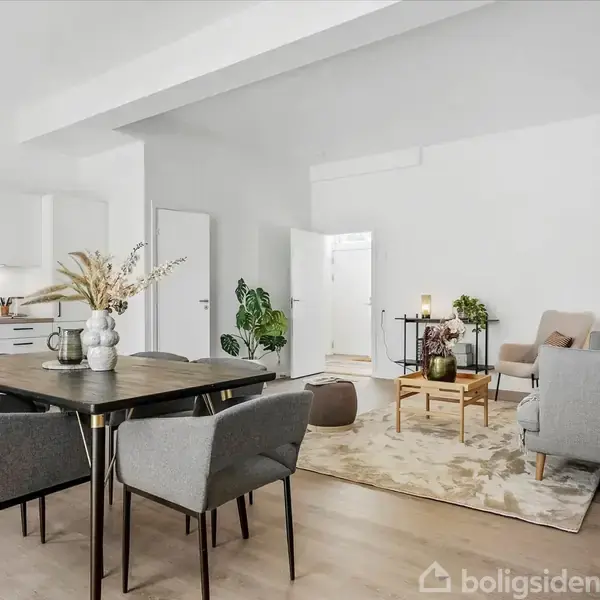 En lys stue med en grå sofa ved et skråvindue, omgivet af en gulvvaseplante og en beige lænestol. En reol til venstre og et mørkt tæppe dækker gulvet.