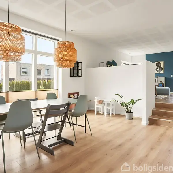 Seng i et lyst soveværelse med sengegavl dekoration. Til venstre ses åben garderobe med tøj. To vinduer giver udsigt til have. Plant og lampe fuldender rummet.